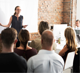A professional white woman stands in front of a diverse group of people in a training class setting.