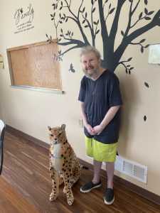 Man in a blue shirt and yellow shorts stands in front of a tree graphic with a stuffed animal cheetah named Tom.