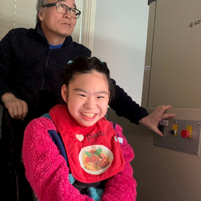 Asian father pushes the buttons of a lift while holding onto the wheelchair with his smiling daughter