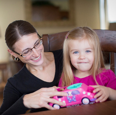 A young mother with dark hair and glasses sits with her blonde toddler daughter at a dining room table