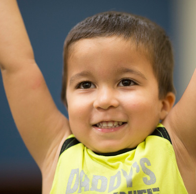 Young Hispanic boy smiles with his arms raised over his head in celebration.