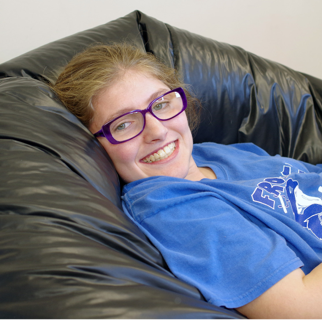 Young white girl with sandy colored hair wearing purple glasses, smiles while leaning back in a black beanbag chair,