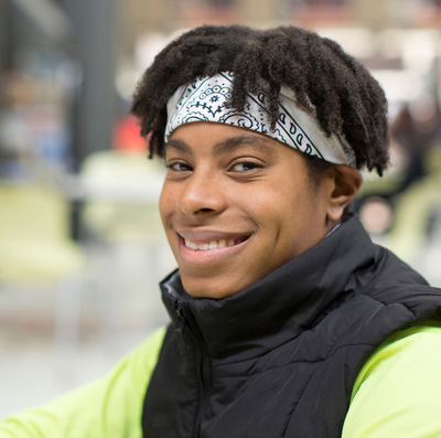 A black student with short dark hair wears a white bandana headband and green shirt with sleeveless vest, smiling and sitting in a school cafeteria.