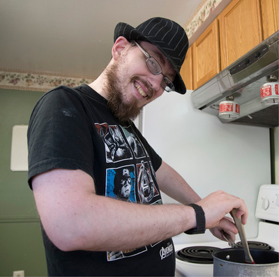 Young man wearing glasses and fedora stands at a kitchen stove stirring soup in a pot.