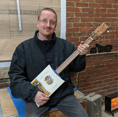 White man with short dark hair wearing glasses holds a guitar created from a cigar box while sitting inside a general store.