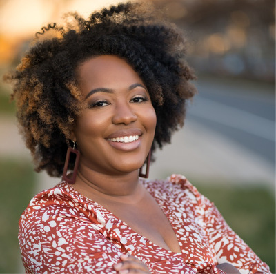 Outdoor portrait of a smiling Black woman with dangling earrings and an orange blouse featuring a white geometric design.