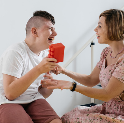 In a home setting, a nurse with brown bobbed hair wearing a floral print outfit assists a young, dark-haired man with a disability who is smiling and holding a large red building block.