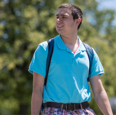 A young man with short dark hair wearing a blue backpack and light blue shirt smiles and walks across a school campus on a sunny day,