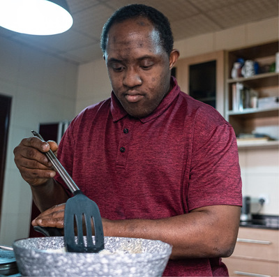 Black man with Down syndrome wearing a red polo shirt attends to cooking food in a stovetop frying pan.