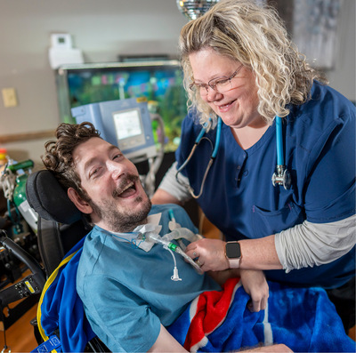 A dark-haired man sitting into a wheelchair smiles while a blonde female nurse wearing navy scrubs and a stethoscope grins down at him and tends to a white medical tube in his neck.