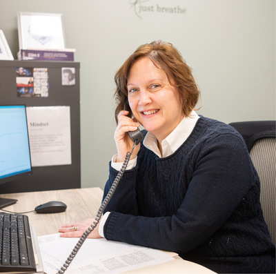 A woman with bobbed red-brown hair sits smiling at an office desk, holding a cord phone to her ear.