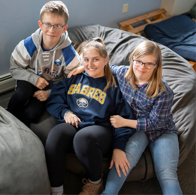 Respite program participants--a young blond boy with glasses and a blonde girl with glasses sit on either side of a female staff member, all smiling and sitting on a beanbag chair.