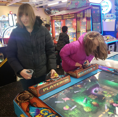 Two young women with light brown hair in an arcade setting, playing a colorful game with joysticks.