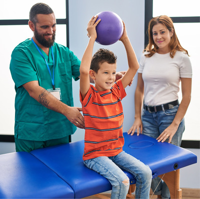 A young boy in an orange striped polo shirt sits on a therapy bench lifting a purple ball over his head, supported by a smiling male therapist in dark aquamarine scrubs while the boy's mother looks on, smiling.