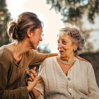 Older woman with grey curly hair sits on a couch, smiling and holding hands with a younger woman whose back is to the camera.