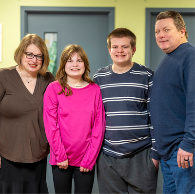 Smiling family of four stands together at a People Inc. program location with pale green walls and grey-blue doors.