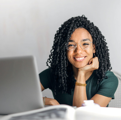 Young Black woman with long, curly dark hair sits at a laptop, smiling.