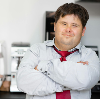 Young man with Down syndrome wearing a button-down shirt and red tie smiles with his arms crossed against his chest.