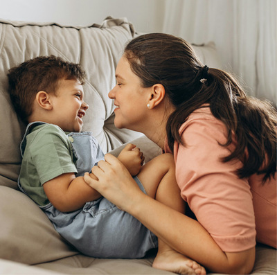 Young boy sitting on a pale beige sofa laughs as his mother leans close to him, smiling.