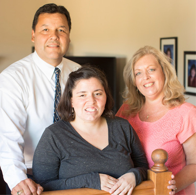 Dark haired young woman smiles and leans against an indoor banister, flanked by a smiling mother on the right and a smiling father on the left.