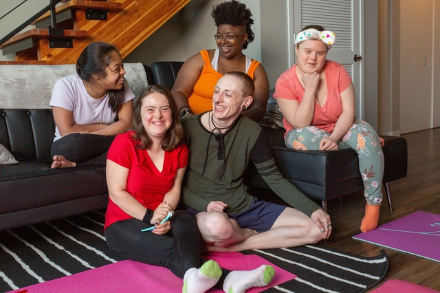 A diverse group of five friends with developmental disabilities sitting together in a living room space, smiling and laughing.