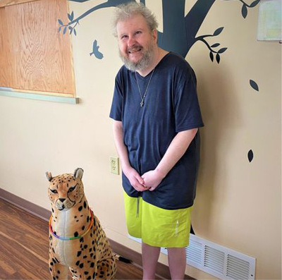 Smiling man stands against a dining room wall featuring a mural of a tree; beside him is a large plush cheetah named Tom.