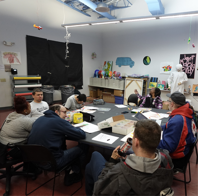 Group of people sit around a table in a classroom style setting, talking and working on art projects.