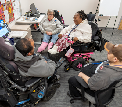 Group of four advocates, including three wheelchair users, meet and converse in a room lined with desks and computer monitors.