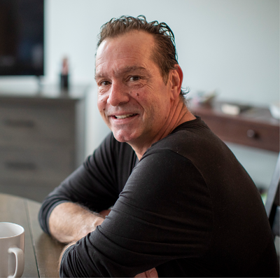 A middle aged man with dark hair sits, smiling, at a round kitchen table.