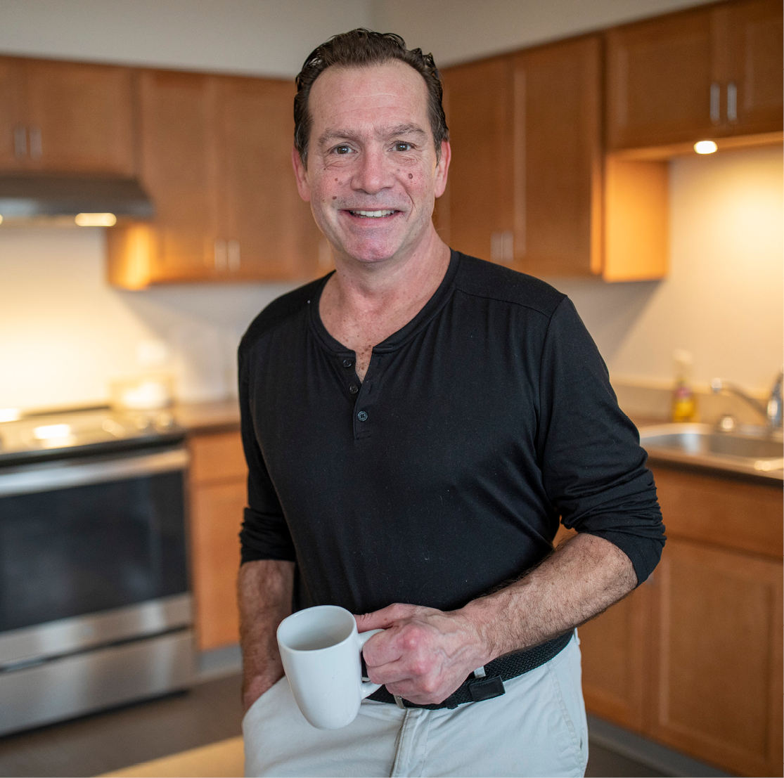 John, a middle aged man with dark hair, stands in a brightly lit kitchen holding a white mug.