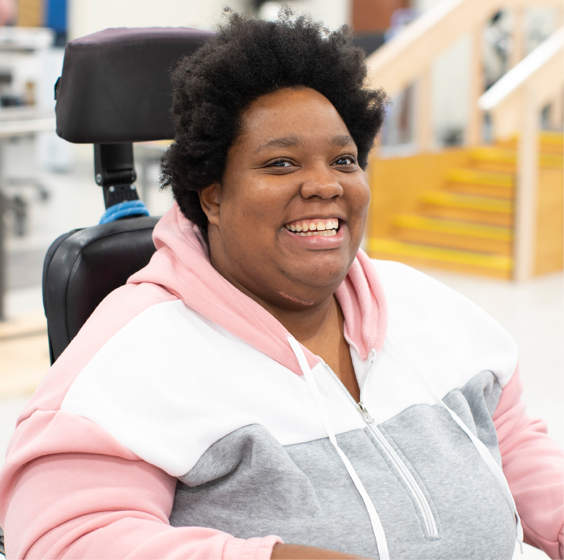 A young Black woman sits, smiling, in a wheelchair in a rehab space.