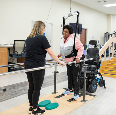 A young Black woman works with a physical therapist to stand out of her wheelchair and walk with the help of grab bars and a support harness.