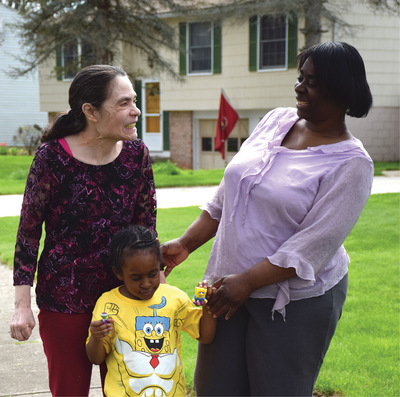 A Perfect Fit participant, a middle-aged brunette woman stands and smiles outside of a house, facing a smiling woman and young child.