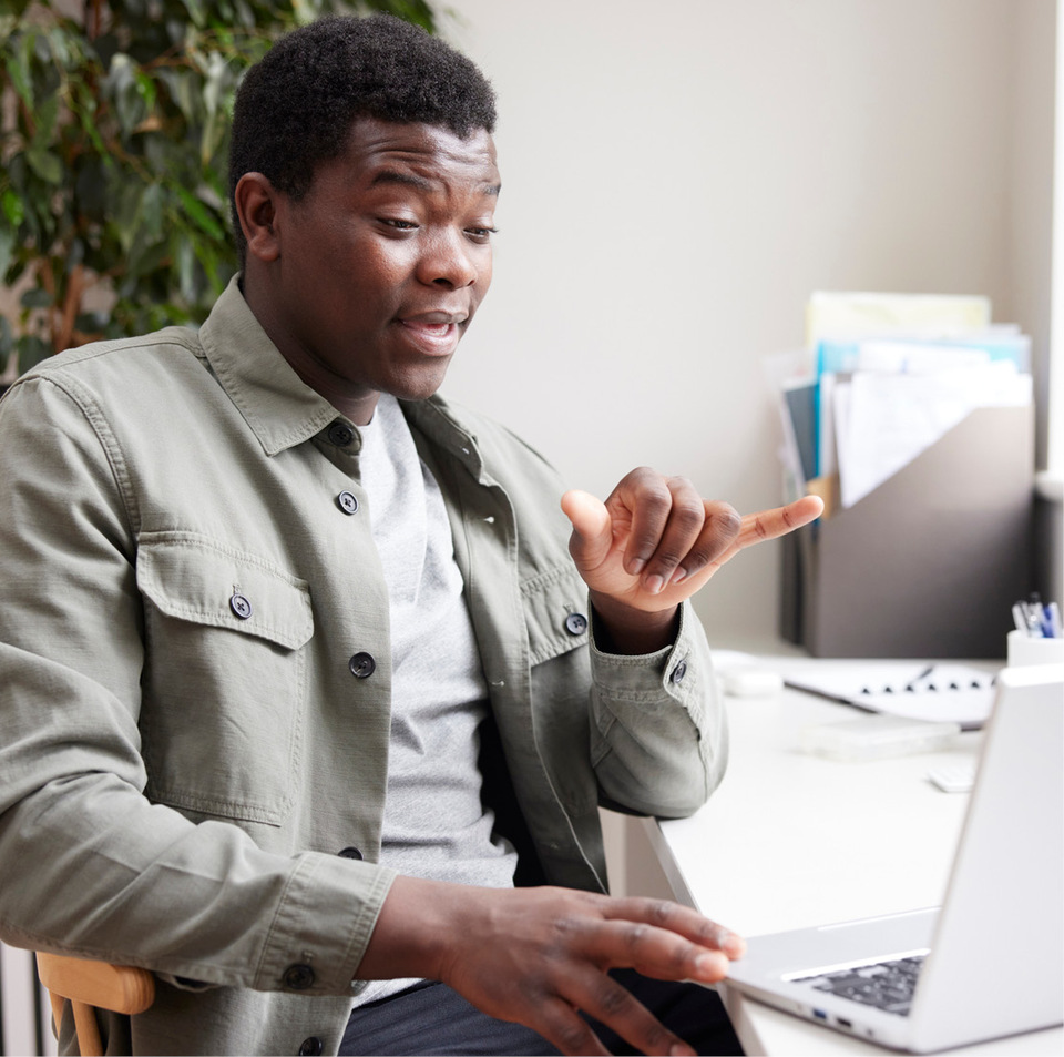 Black man sits at a table facing an open laptop, communicating through sign language.