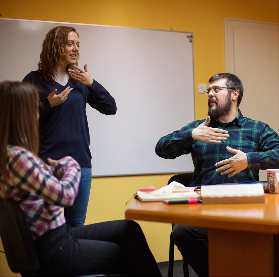 A young man and woman sit at a classroom table practicing sign language while a female instructor stands signing nearby.