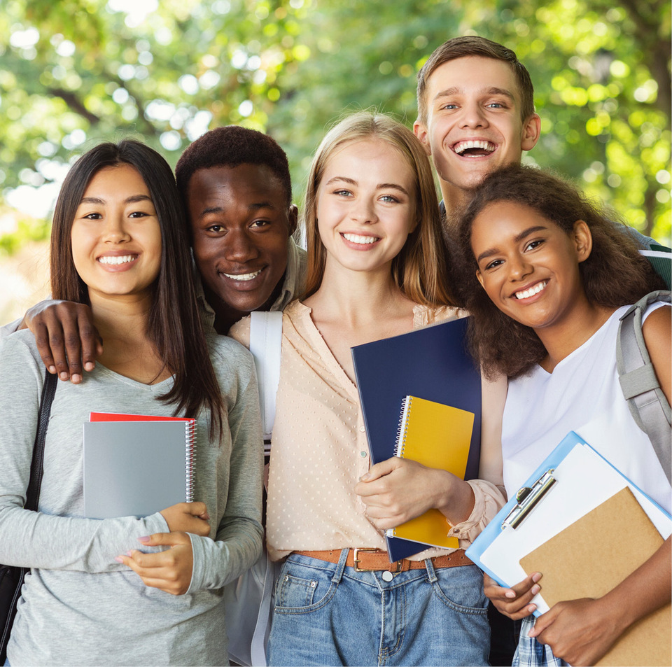 A group of diverse students stand outside on a sunny day, smiling and holding an assortment of folders and notebooks.
