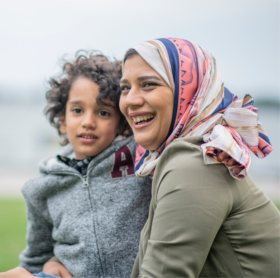 Smiling immigrant woman wearing a colorful head scarf holds her young son, backed by an outdoor lake landscape.