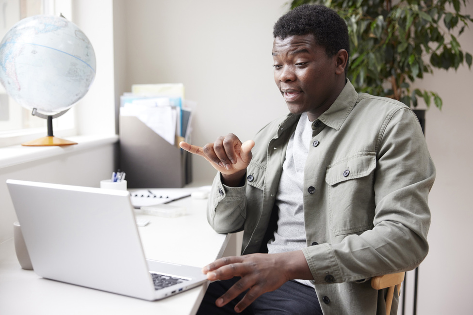 Black man sits at a table facing an open laptop, communicating through sign language.