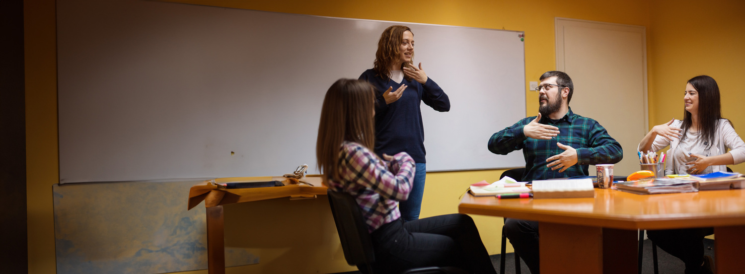 A group of ASL practice communicating in American Sign Language while an instructor stands nearby in front of a white board.