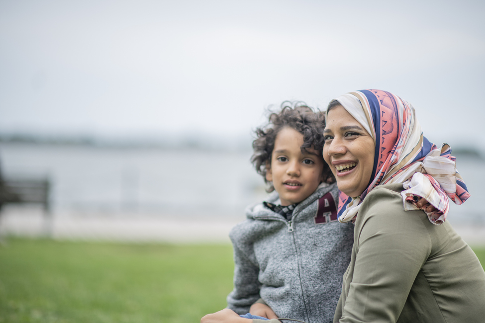 Refugee woman wearing a colorful headscarf smiles while holding her young son and standing in front of a lakeside landscape.