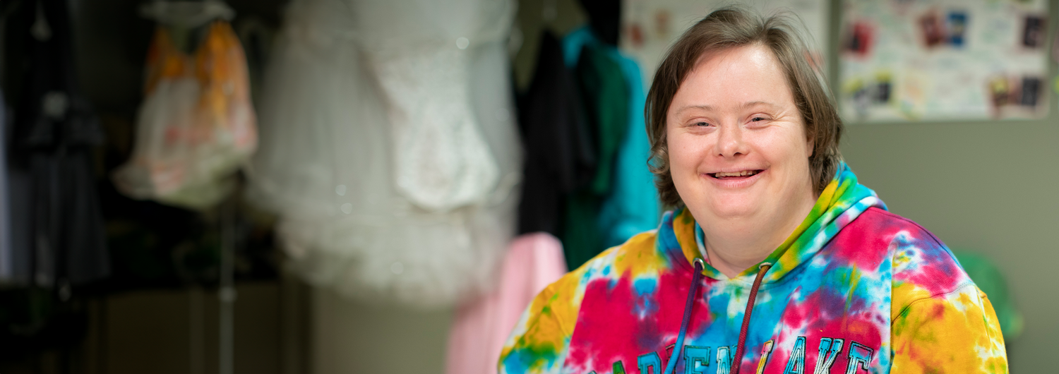 Shop Help ensure that people with developmental disabilities, special needs and older adults receive the supports they need. A young woman with Down syndrome smiles, wearing a colorful tie-dye hoodie