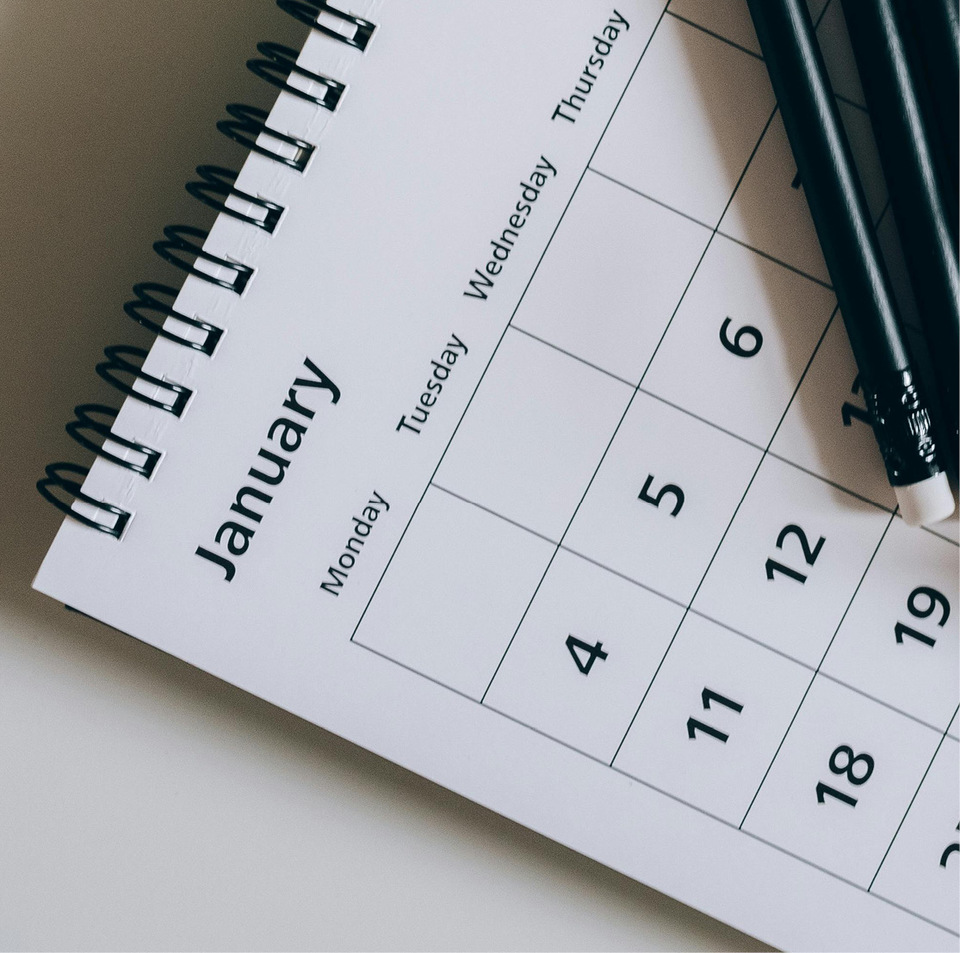 A blank white spiral calendar, open to the month of January with a pair of black pencils laying on top.