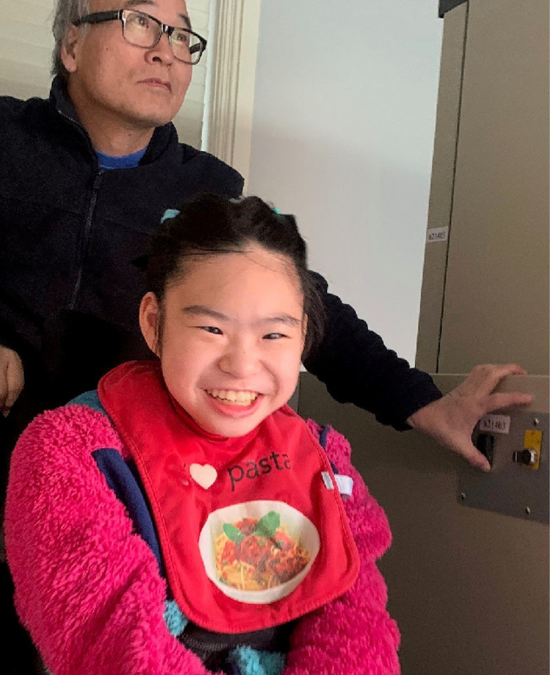 Asian man presses a wheelchair lift button to assist his 14-year old daughter, who is smiling and seated in front of him.