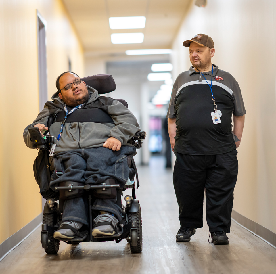 Two advocates, one young man in a wheelchair beside another young man, move together down a cream-colored hallway.