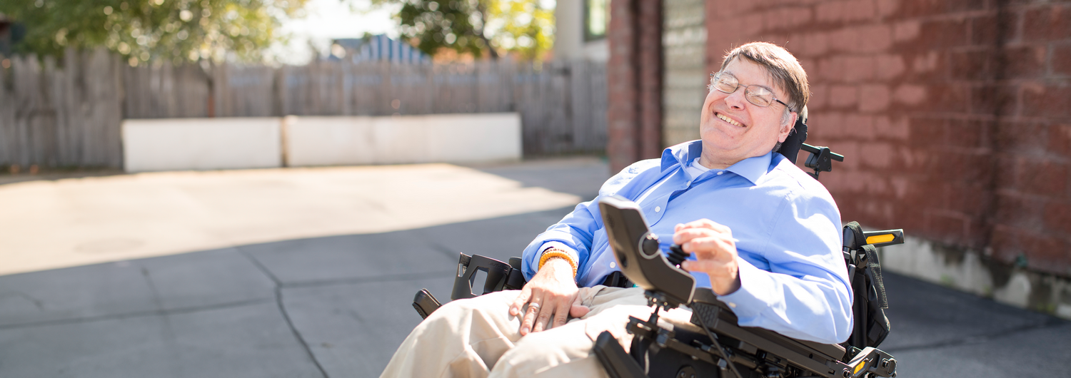 Man wearing glasses wears a big smile as he sits outside on a sunny day in his wheelchair.